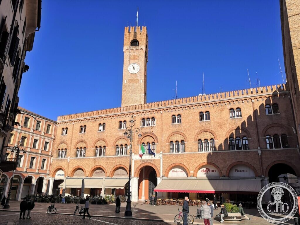 A long brick palace with crenellations and arcades on a sunny square, a tall brick tower with a clock rising above it