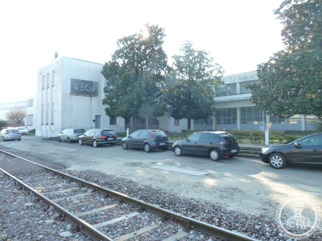 View along a railway siding towards a white block with the VEGA sign and a long glazed building behind trees