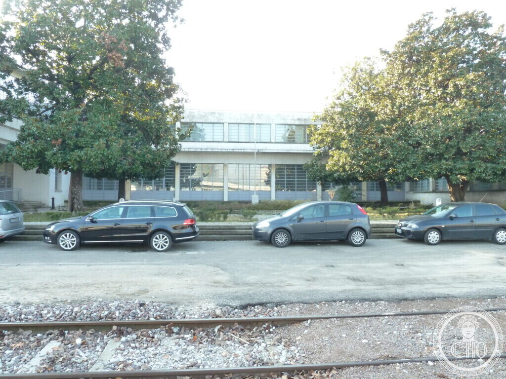 A long low white building with bands of steel-framed windows behind two large magnolia trees, parked cars and rails in front