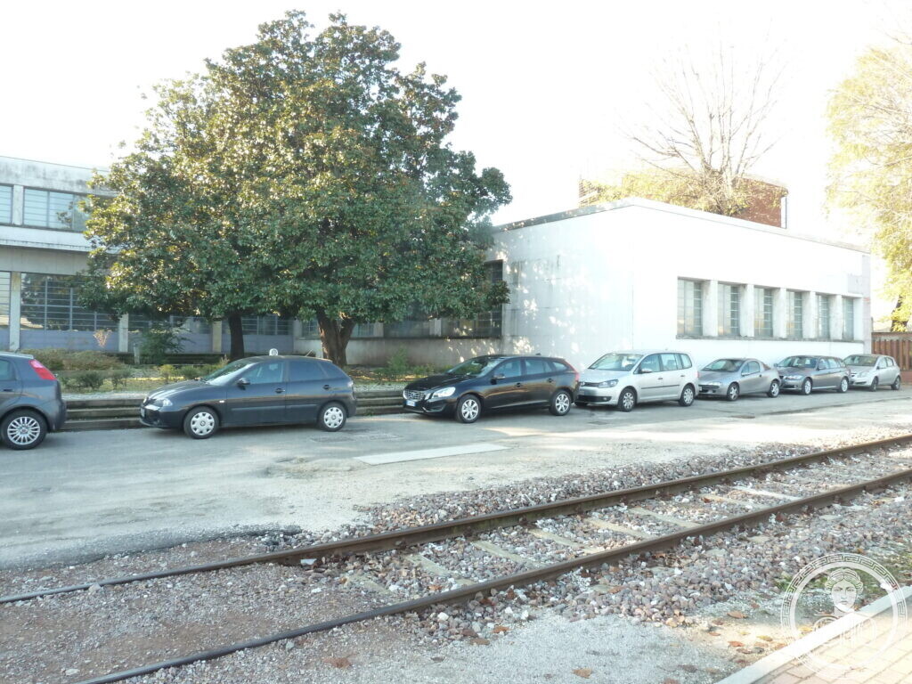 A low white building with a row of square windows next to a larger glazed block, cars parked along a railway track