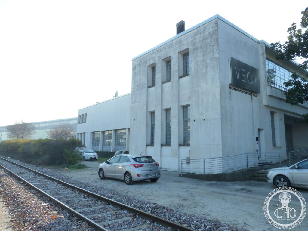 A tall white rendered building with narrow vertical windows and the VEGA sign on its corner, a railway track in the foreground