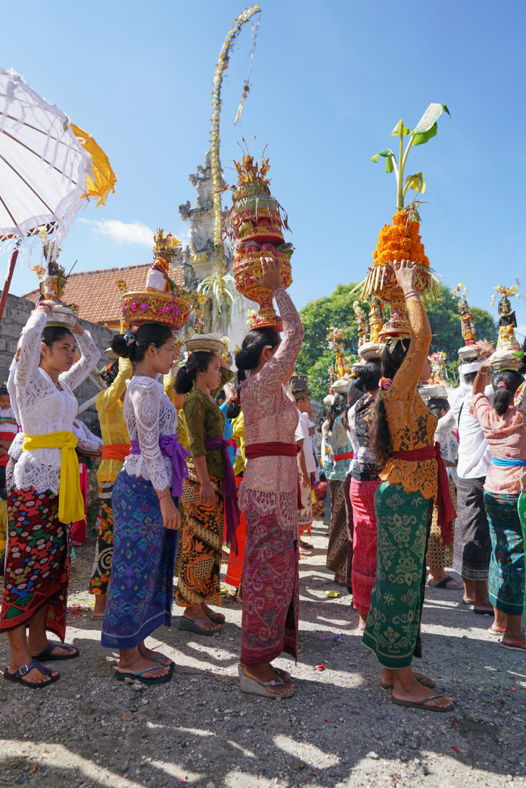 A procession of Balinese women in ceremonial dress carrying offerings on their heads along a road, temple umbrellas and tall offerings behind