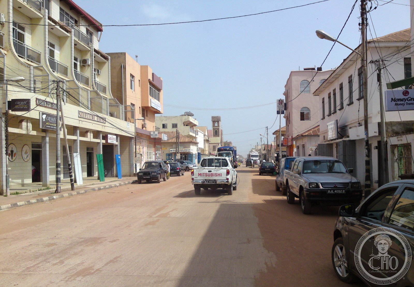 A busy unpaved street in Banjul, The Gambia, with the square brick bell tower of the Cathedral of Our Lady of the Assumption visible above the rooftops