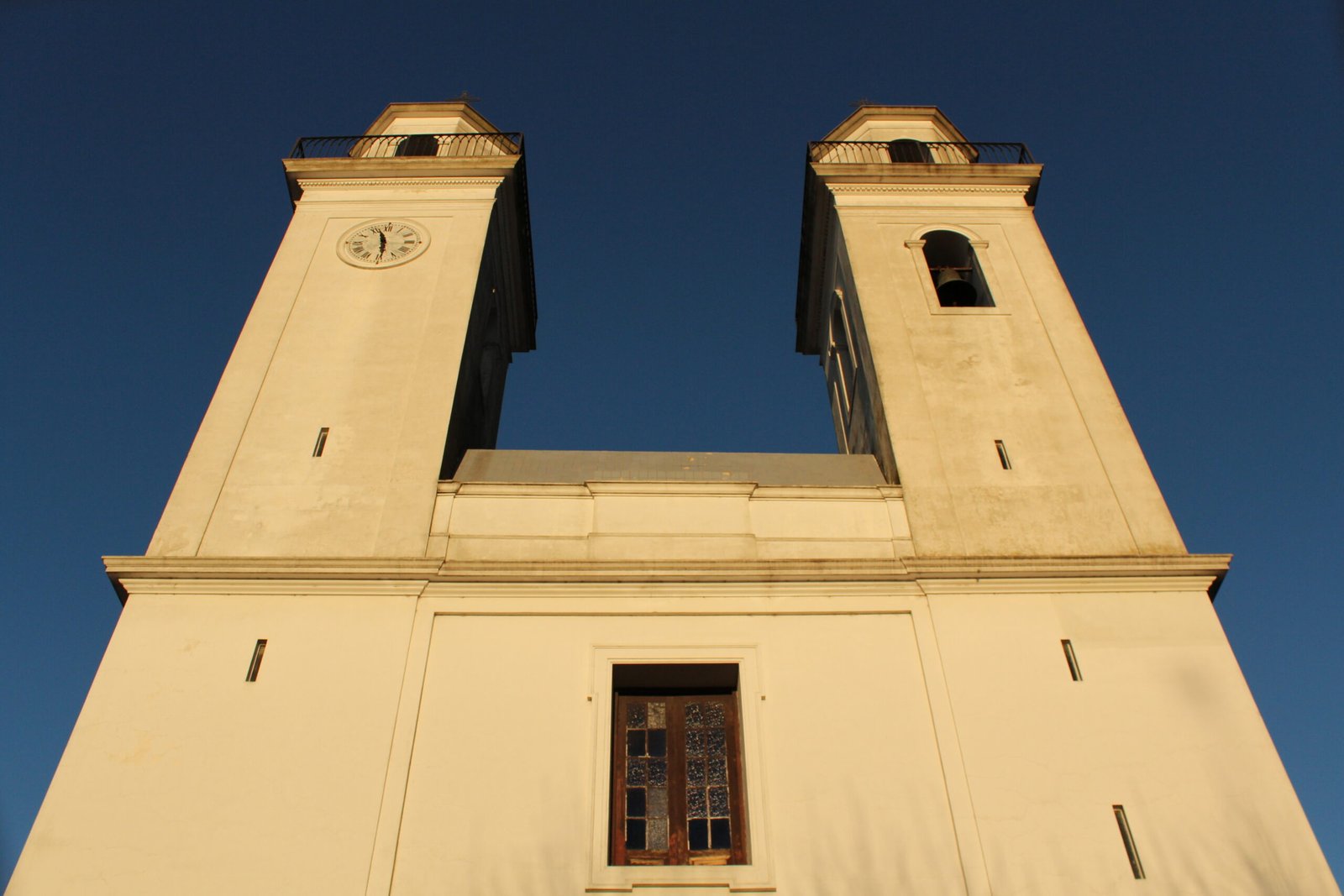 The twin white bell towers of the Basilica of the Holy Sacrament in Colonia del Sacramento, one with a clock and one with a bell, against a blue sky