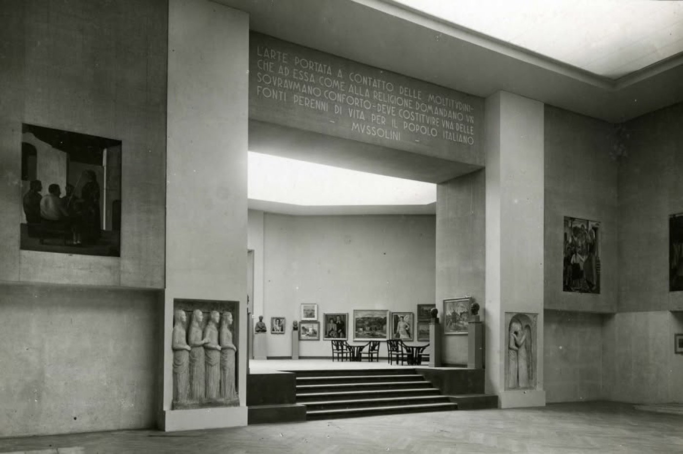 Hall of a 1930s art exhibition with paintings and reliefs and, above a monumental opening, an inscription attributed to Mussolini on the function of art