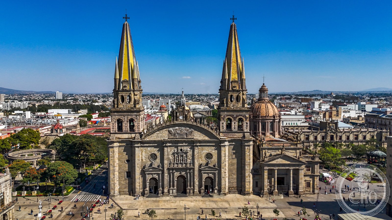 Aerial view of Guadalajara Cathedral, Mexico, showing its twin neo-Gothic spires covered in yellow tile, a rose window, and the Plaza de Armas below