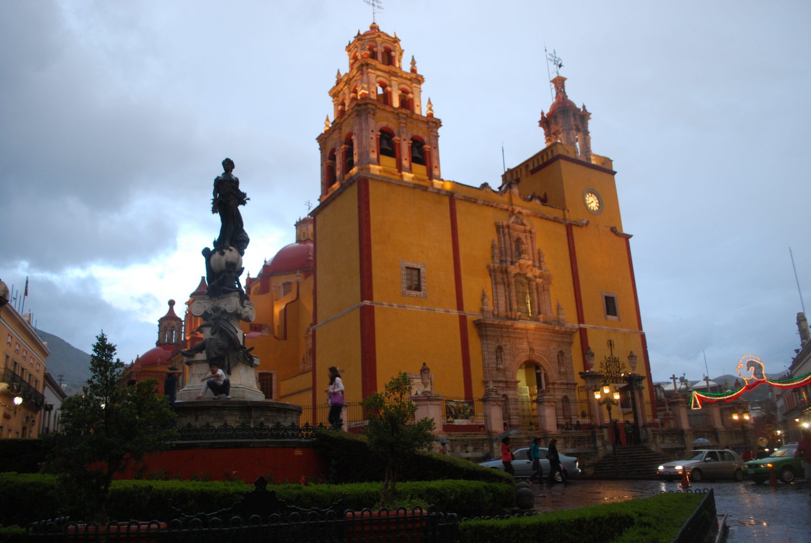 The ochre-yellow Baroque facade of the Basilica of Our Lady of Guanajuato with its twin asymmetric towers, floodlit at dusk on the Plaza de la Paz