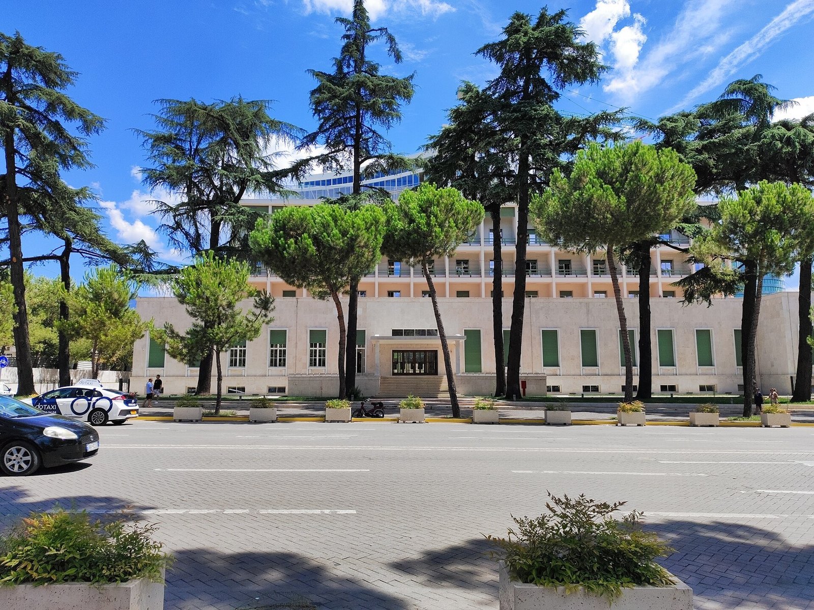 The long white Rationalist facade of the former Hotel Dajti in Tirana, seen through pine trees