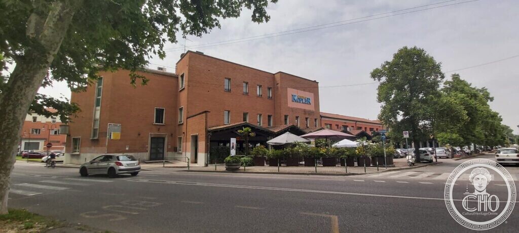 A long brick industrial building with a Kofler sign on its front and a restaurant terrace with umbrellas at street level