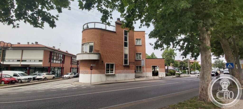 A brick corner building with a rounded end, a balcony, a narrow vertical window and a metal railing on the roof, plane trees in front