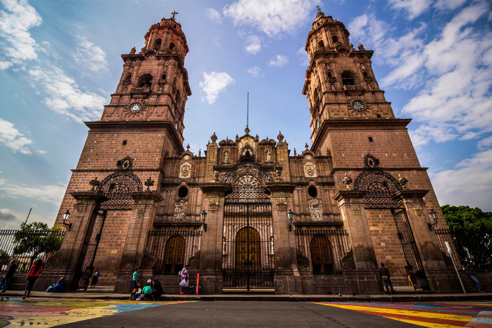 Front view of Morelia Cathedral, Mexico, with twin pink-stone baroque towers, clocks, and wrought-iron atrium gates
