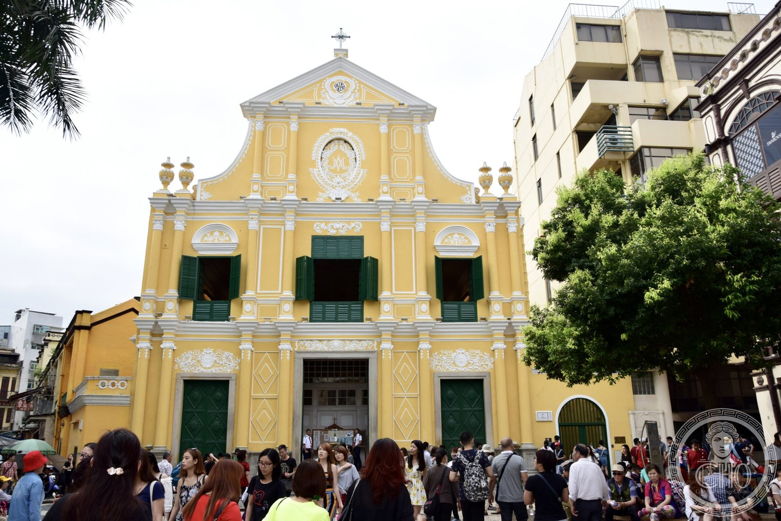 St. Dominic's Church, Macau, a pale-yellow baroque facade with white stucco ornament, green-shuttered windows, a cross atop the pediment, seen from Senado Square
