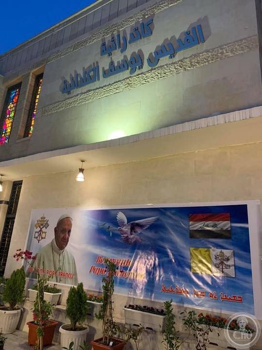 St. Joseph Chaldean Cathedral in the Karrada district of Baghdad, Iraq, at dusk, with a Pope Francis welcome banner and Arabic signage above the entrance