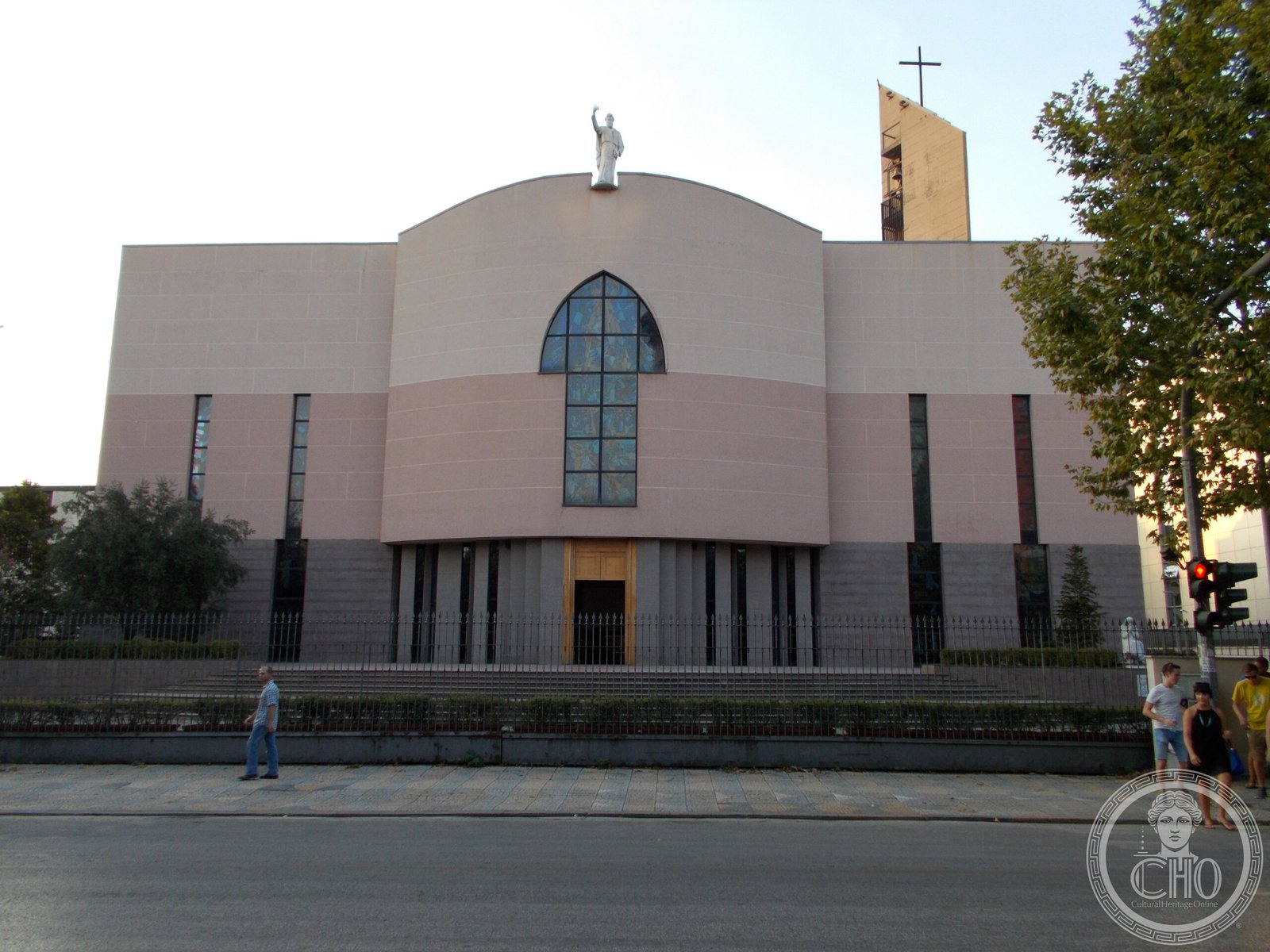 St. Paul's Cathedral in Tirana, Albania, a modern salmon-pink stone facade with a tall pointed-arch stained-glass window and a statue of St. Paul on the roofline