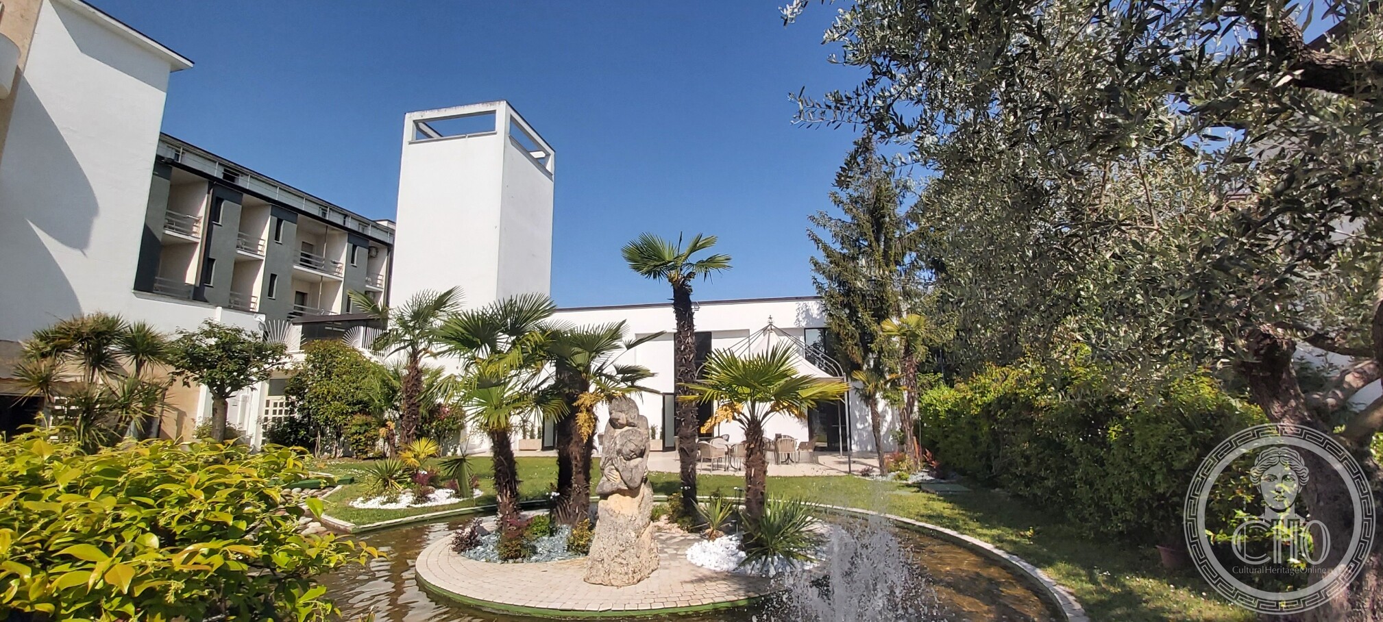 The white tower of the former Casa del Fascio rising above a hotel garden with palms, a fountain and garden furniture