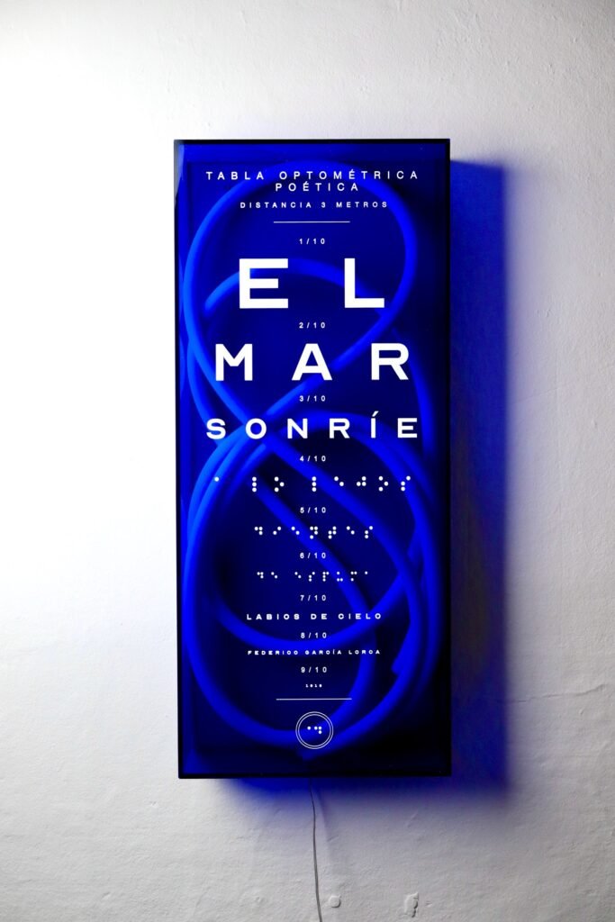 A blue lightbox in the form of an eye chart reading EL MAR SONRIE in descending sizes, with lines of Braille below