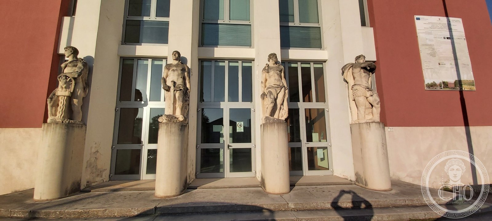Four stone male figures on cylindrical plinths in front of the glazed entrance of the Casa del Balilla, with the house number 37 on the wall