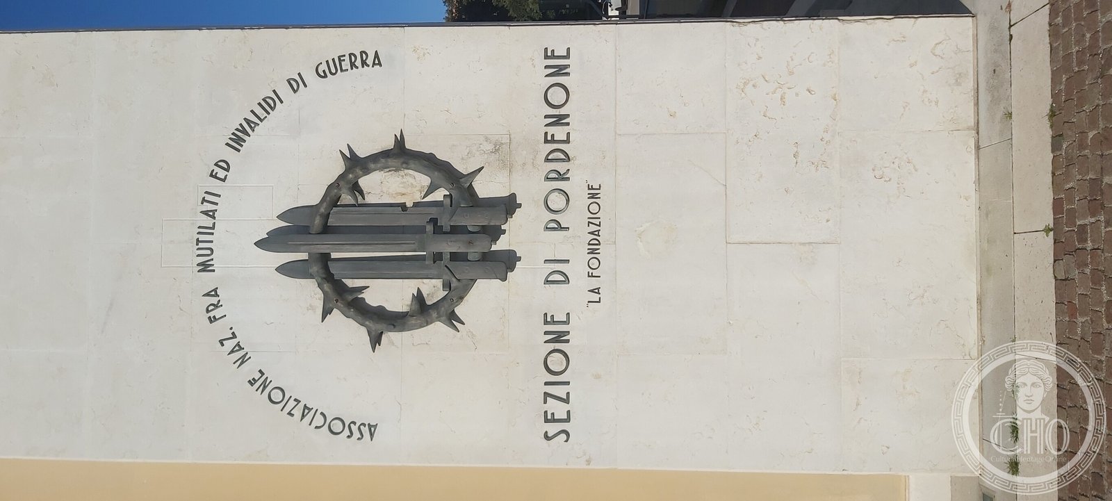 Bronze emblem on the stone pier: a crown of thorns around three crossed swords, ringed by the name of the National Association of War Disabled and Invalids, Pordenone section