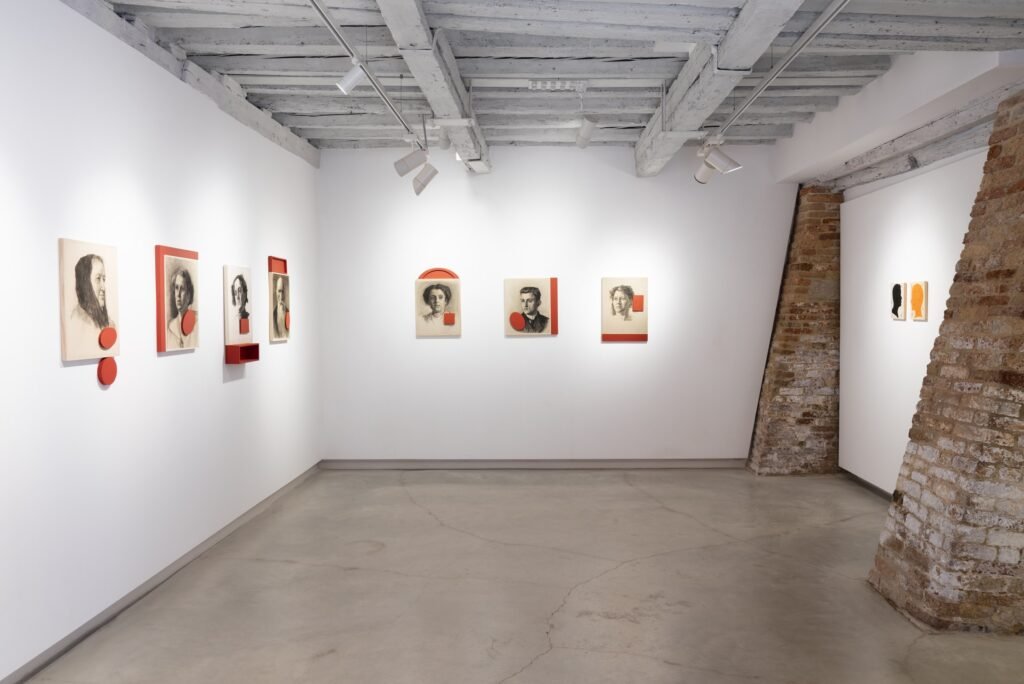 Gallery room with small portraits hung in a line, red discs and panels beside them, and an exposed brick pier at the right