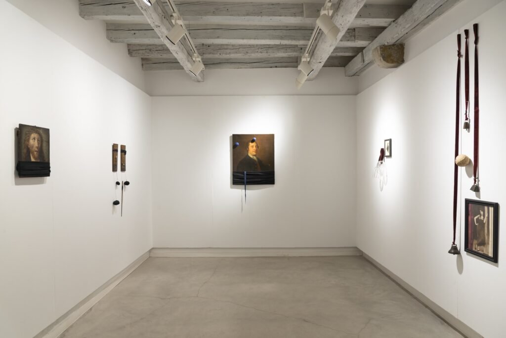 Gallery room with a nineteenth-century male portrait bound with dark cloth, a small framed photograph, and leather straps hanging from the wall
