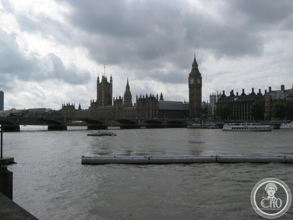 A long Gothic Revival palace along a wide grey river under heavy cloud, a bridge in front of it and a tall clock tower at its right end