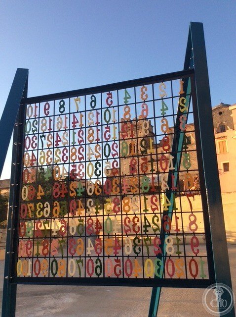 The multiplication table of the sculpture seen from behind, with the stone buildings of Stilo visible through the grid