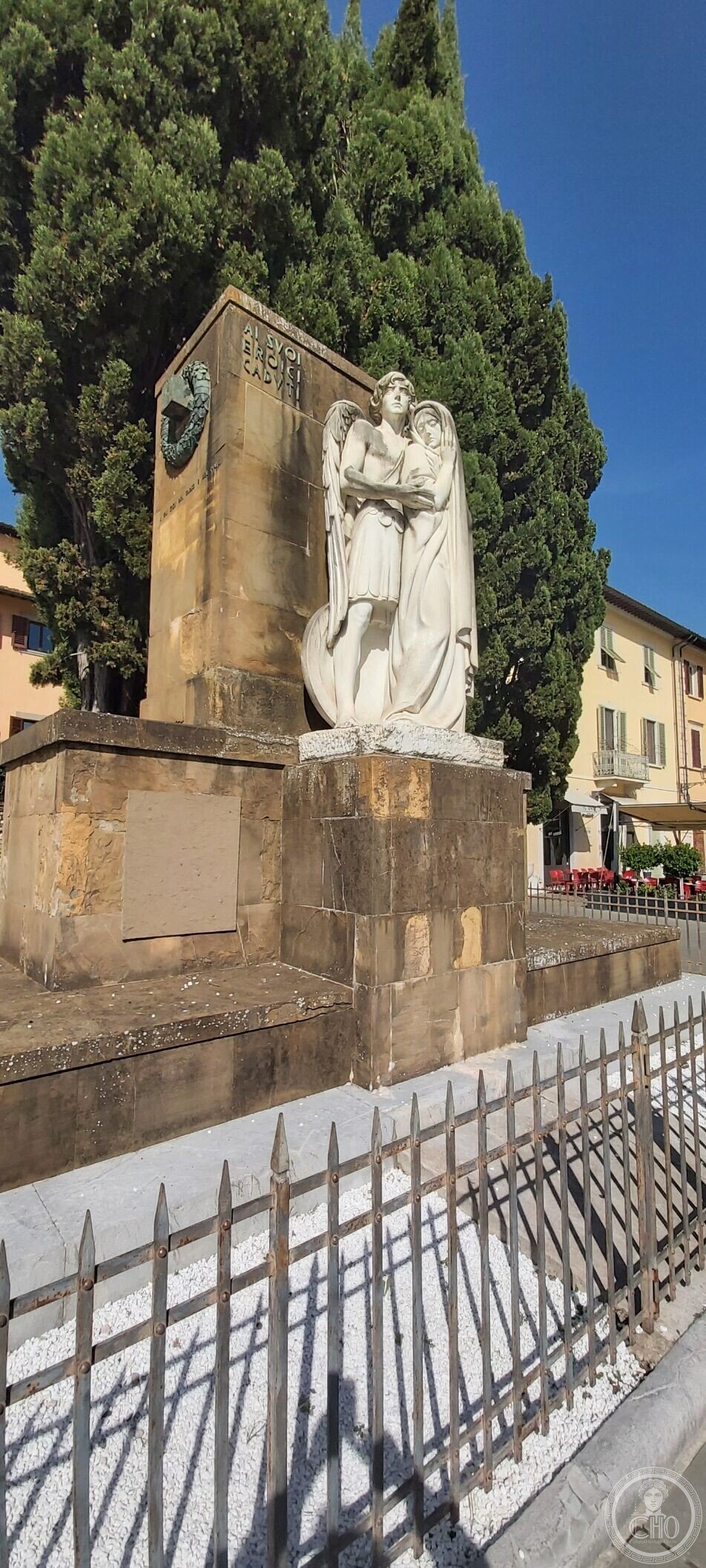 Closer view of the Prato war memorial group and the bronze shield on the stele, with cypresses behind