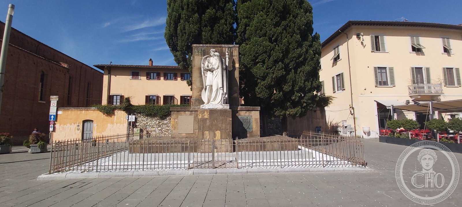 The Prato war memorial seen across the paved Piazza delle Carceri, with the church on the left and cypresses behind