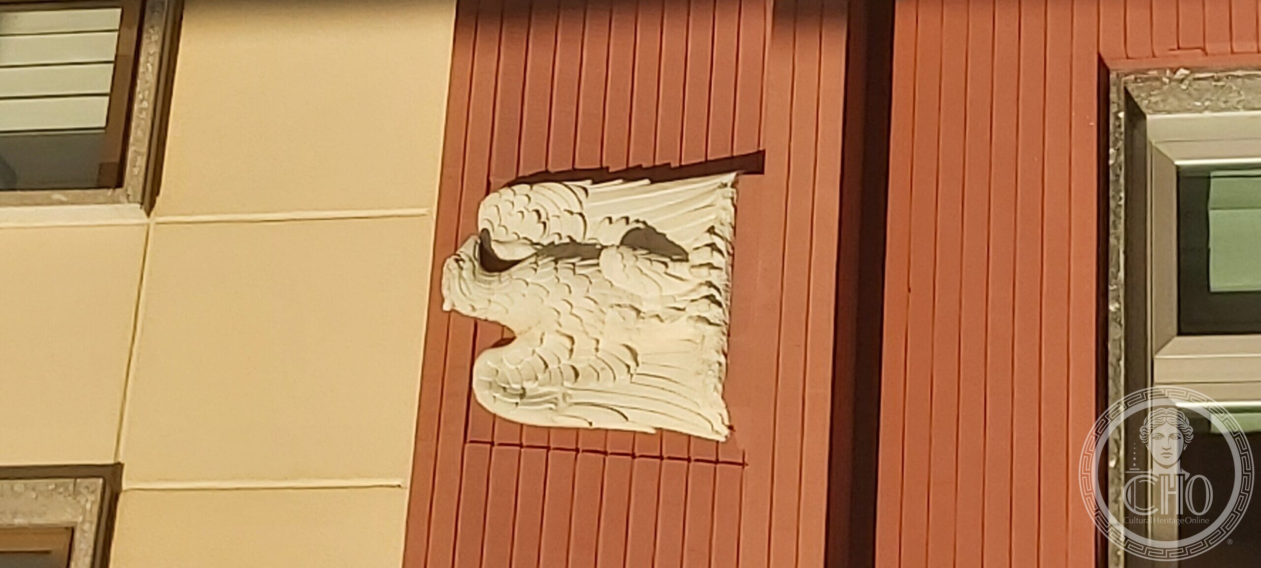 White stone relief of an eagle with spread wings in a square panel, set against the red ribbed Litoceramica facing