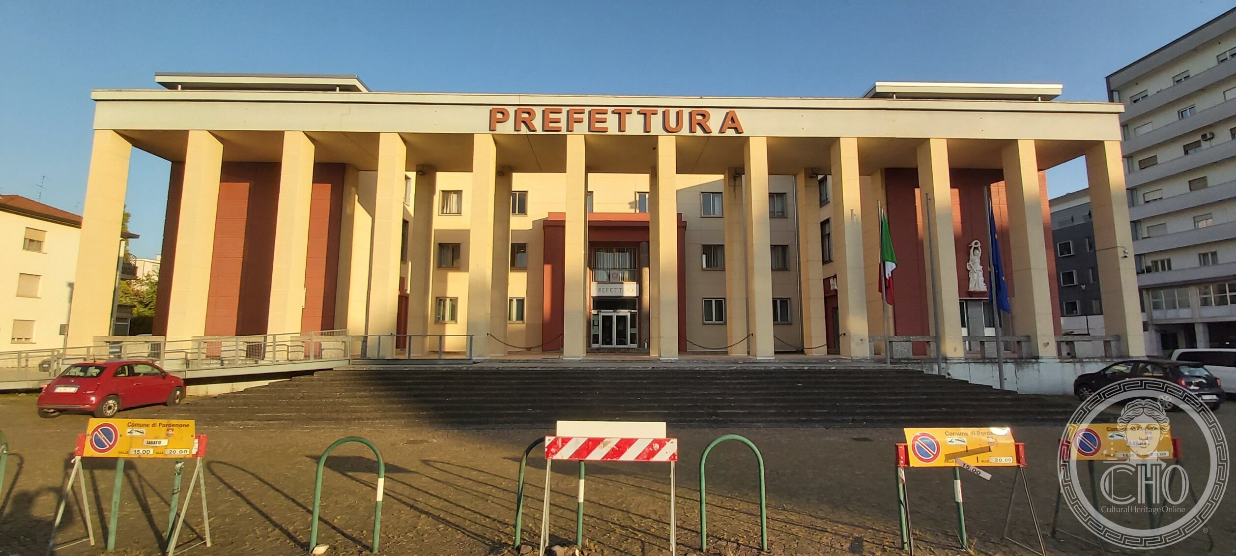 The former Casa del Fascio seen frontally from the square, raised on its podium above a wide flight of steps, with flags at the entrance