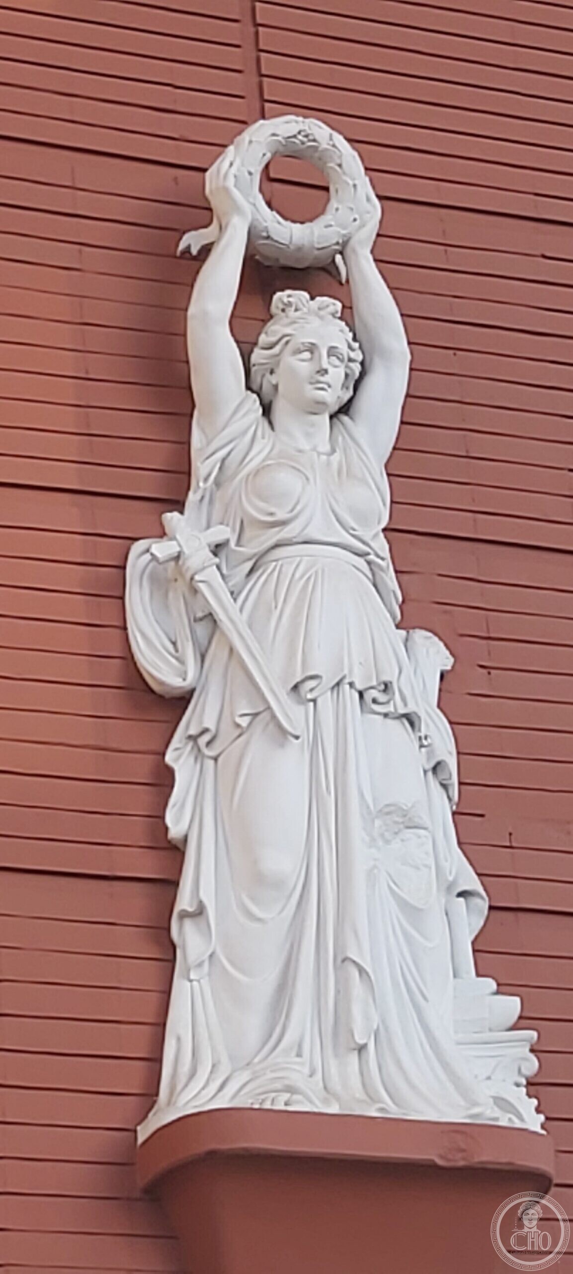 White stone relief of a draped female figure on a bracket, arms raised holding a laurel wreath, a sword at her side, against red ribbed facing