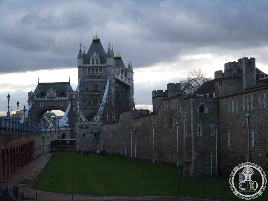 Two stone towers of a bridge with green roofs rising beyond the battlemented curtain wall and grassed moat of a medieval castle, under a dark sky