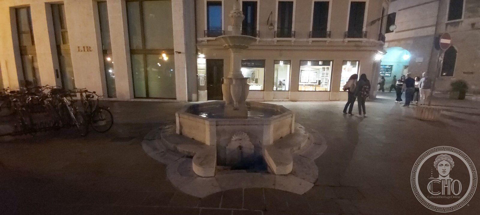 The fountain of Piazza San Vito seen across the paved square in the evening, with shop fronts and passers-by behind