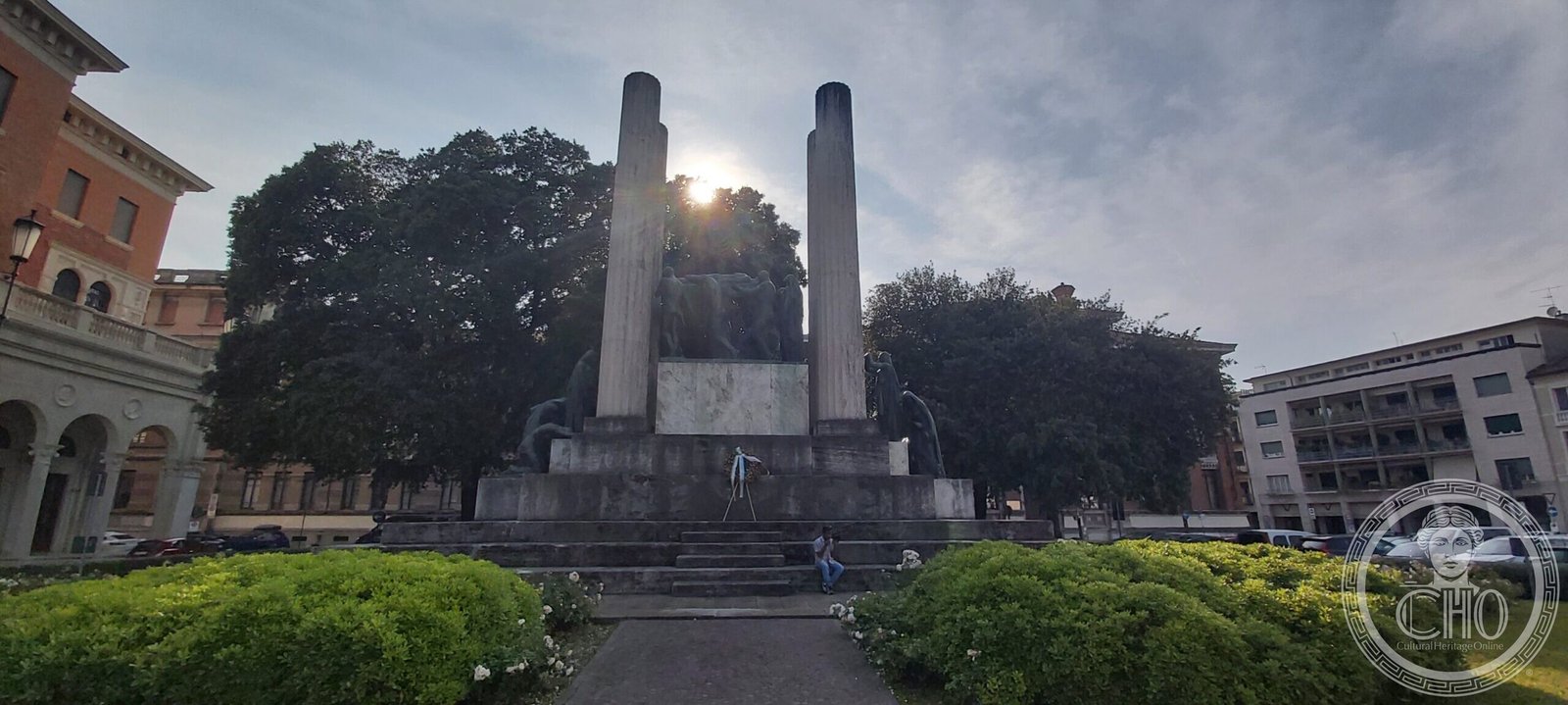 The war memorial of Treviso: two tall marble pylons with bronze figures on stepped platforms between them, seen against the sun
