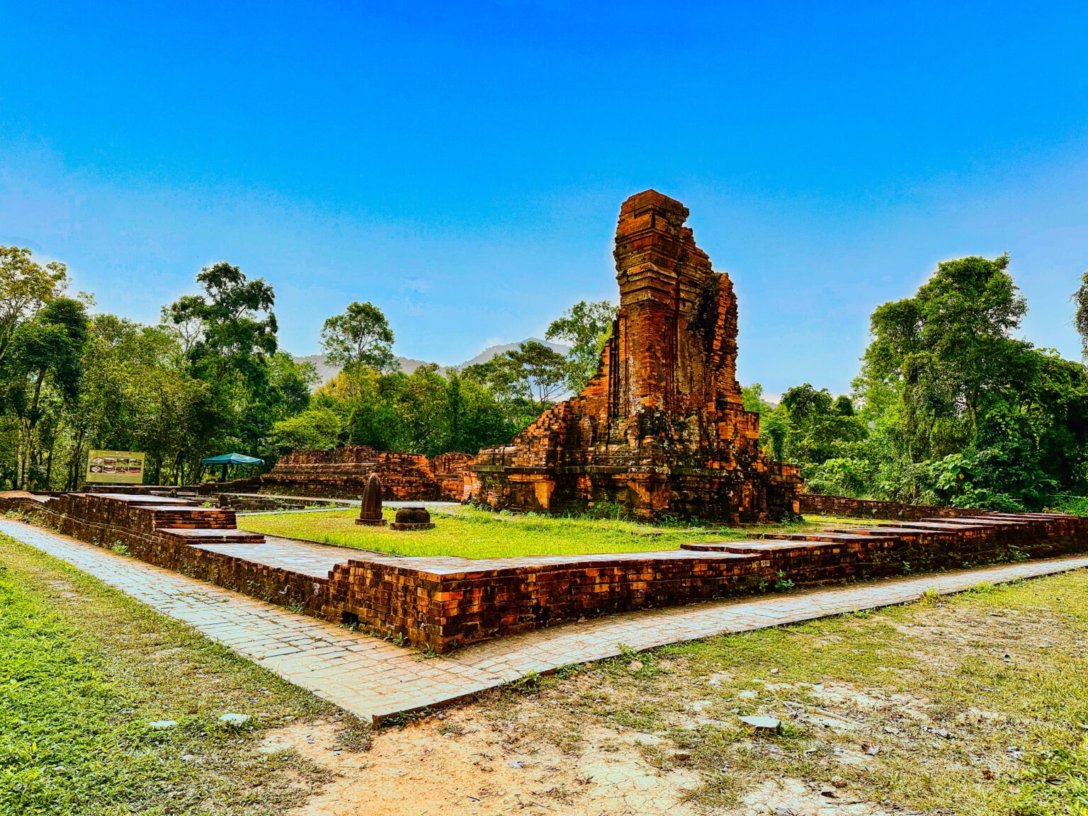 A single broken brick tower standing on a cleared brick platform at Mỹ Sơn, the rest of the group gone, forest behind