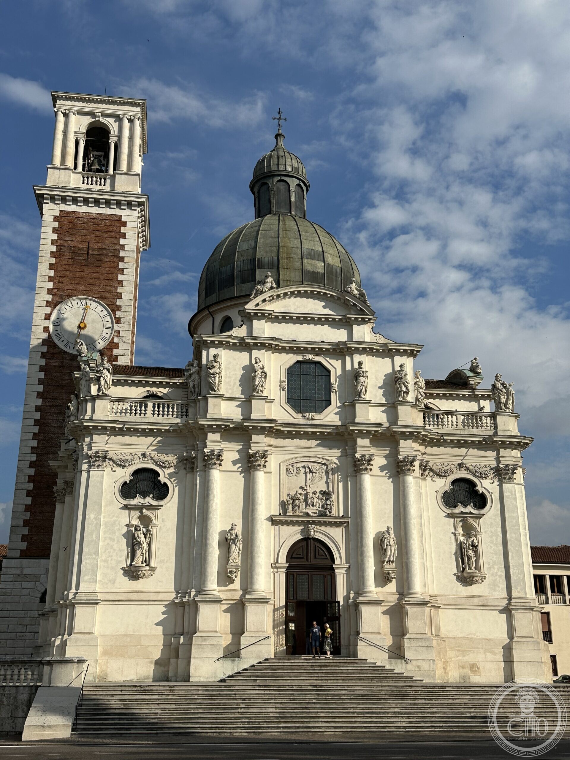 The Baroque facade of the Basilica di Monte Berico in Vicenza, with its bell tower and green dome, seen from the steps below