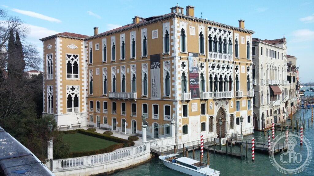 The ochre Gothic front of Palazzo Cavalli-Franchetti on the Grand Canal in Venice, its white traceried windows in two tiers, with a walled garden at the left and mooring poles in the water