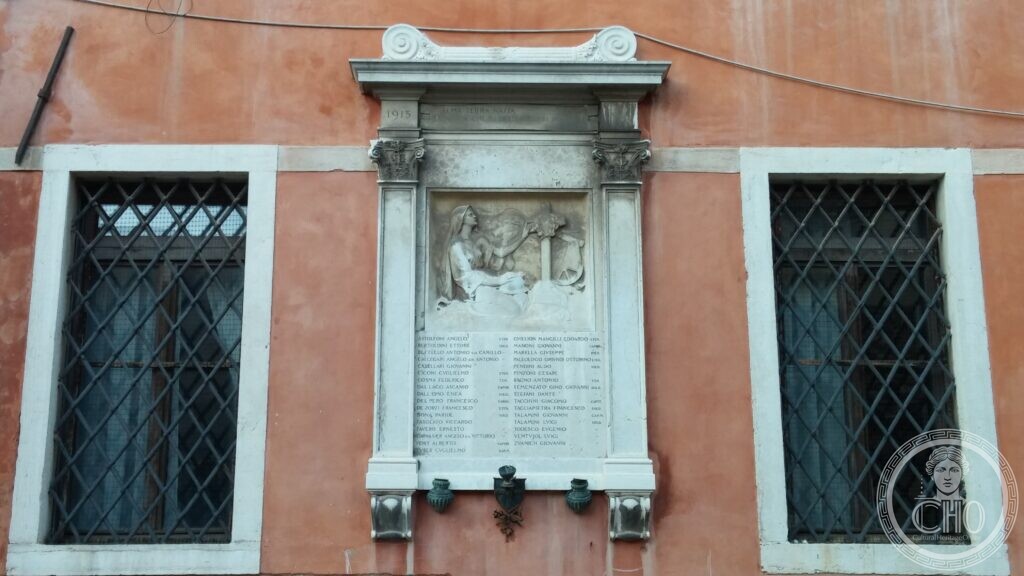 A marble war memorial framed by two Corinthian columns and a pediment on a red plastered wall, a carved relief of a winged Victory above the names of the fallen, two bronze lamps below