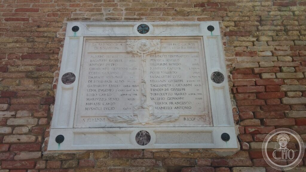 A white marble war memorial set in a brick wall, framed in Istrian stone with dark red roundels at the corners and bronze plaques, the names of the fallen cut in two columns