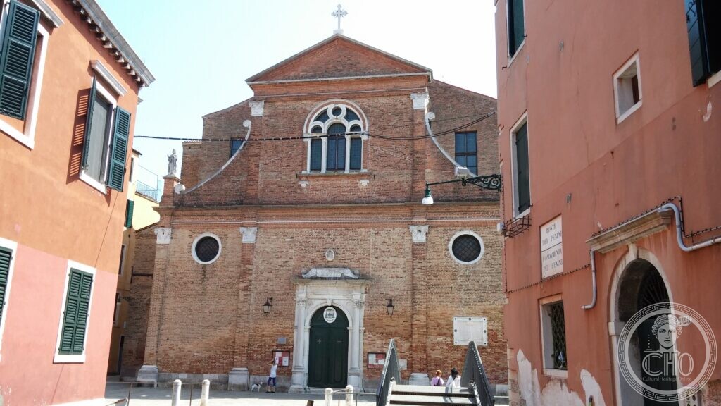 The brick front of the church of San Martino in Venice, with a trefoil Gothic window above the stone portal and two round windows at the sides, seen across the campo