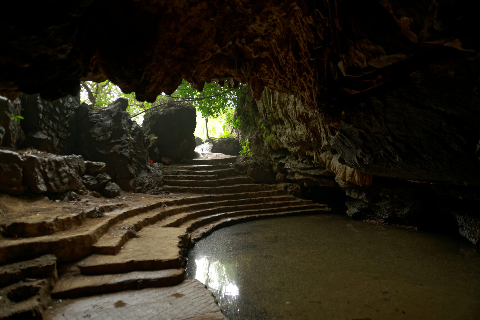 The mouth of a cave at Trang An opening onto green light, with worn stone steps rising from a pool of water inside