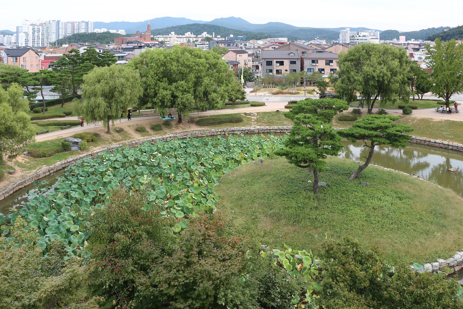 Section of the Hwaseong Fortress wall with a curved stone battlement and watchtower rising above green trees in Suwon