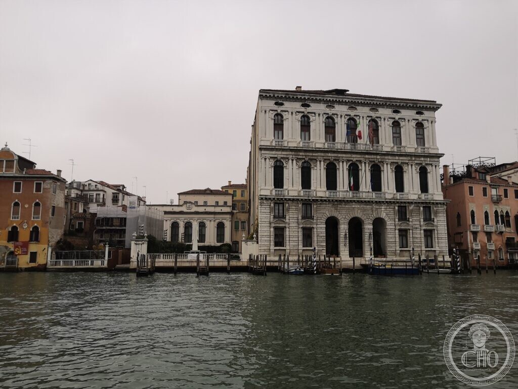 The white stone front of Palazzo Corner della Ca' Granda across the Grand Canal in Venice, three arched water gates on a rusticated ground floor, two upper floors of arched windows between paired columns, Italian flags at the windows