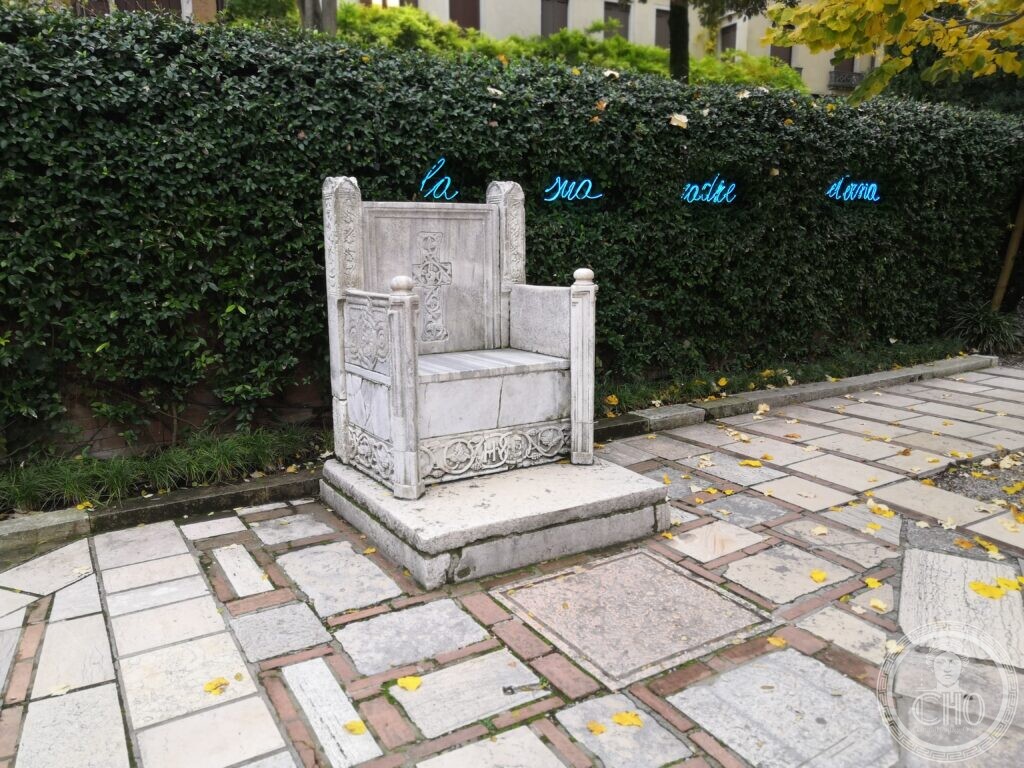 A carved marble throne on a stone platform in the garden of the Peggy Guggenheim Collection, a line of words in blue neon along the ivy-covered wall behind
