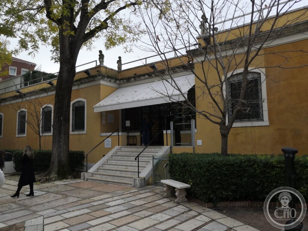 The low ochre wing on the garden side of the Peggy Guggenheim Collection, stone steps up to an entrance under a white awning, trees in the paved garden