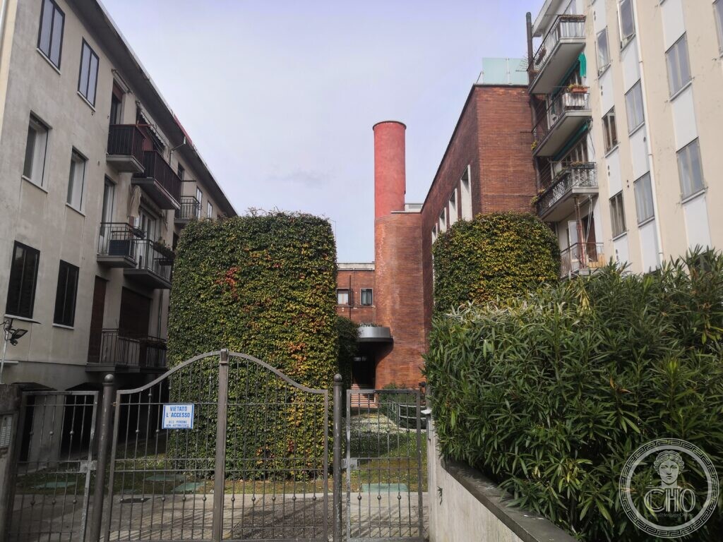 The red-brick former heating plant of Venice railway station, its tall round chimney rising between two clipped hedges, framed by pale postwar apartment blocks, a closed iron gate in front