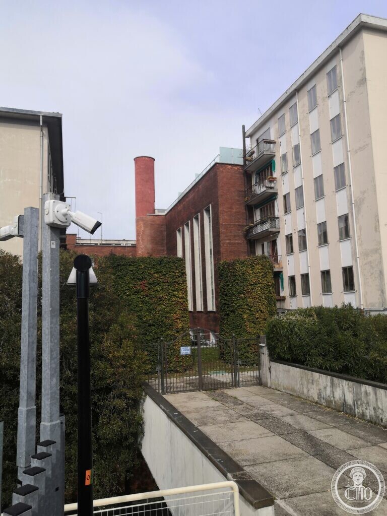 The former heating plant of Venice station seen from a ramp, the round brick chimney and the brick block beyond a gate and hedges, apartment blocks on the right