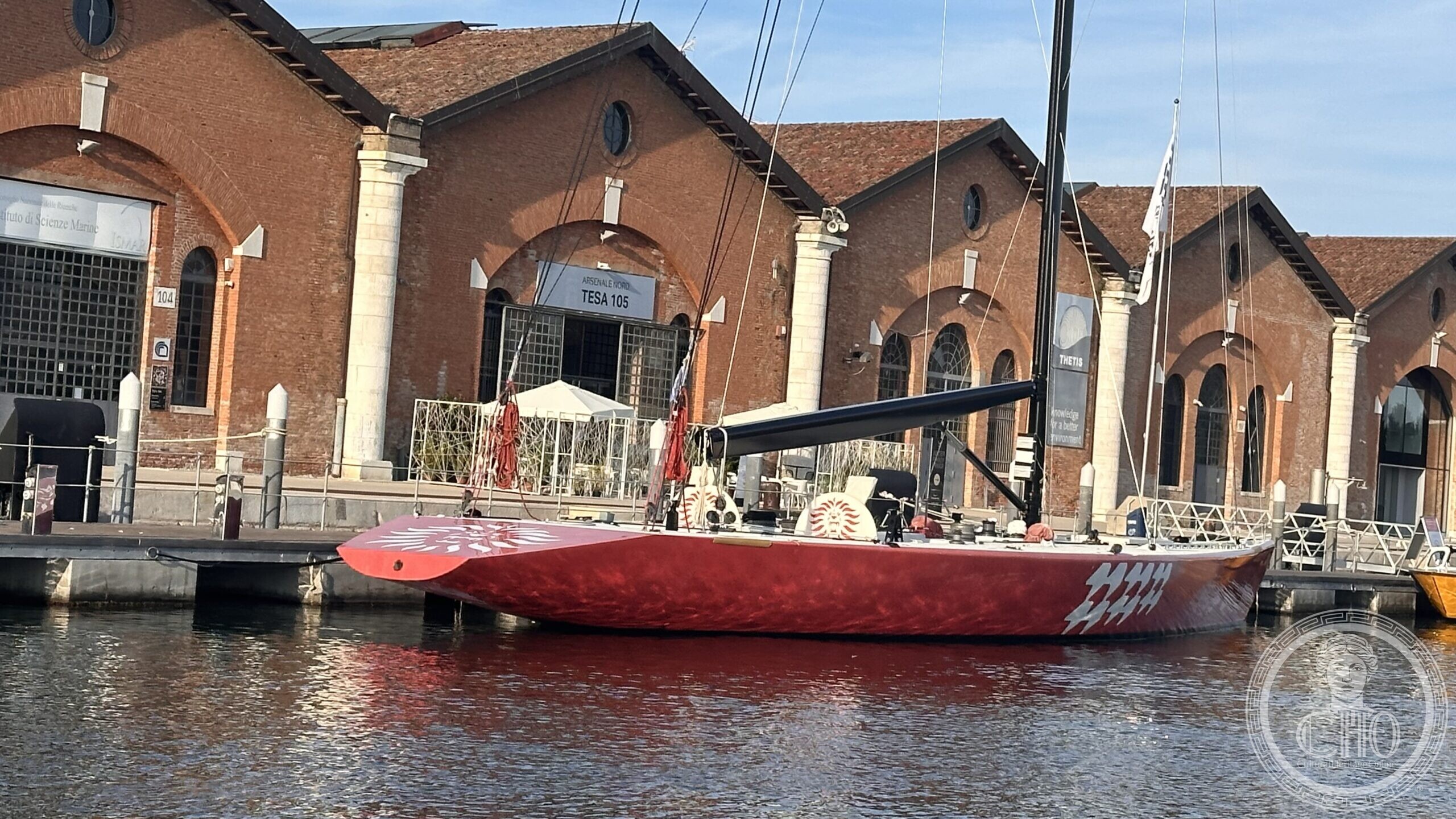 A red-hulled Il Moro di Venezia America's Cup yacht moored at Arsenale Nord, Venice, in front of the brick sheds of the Novissima dock