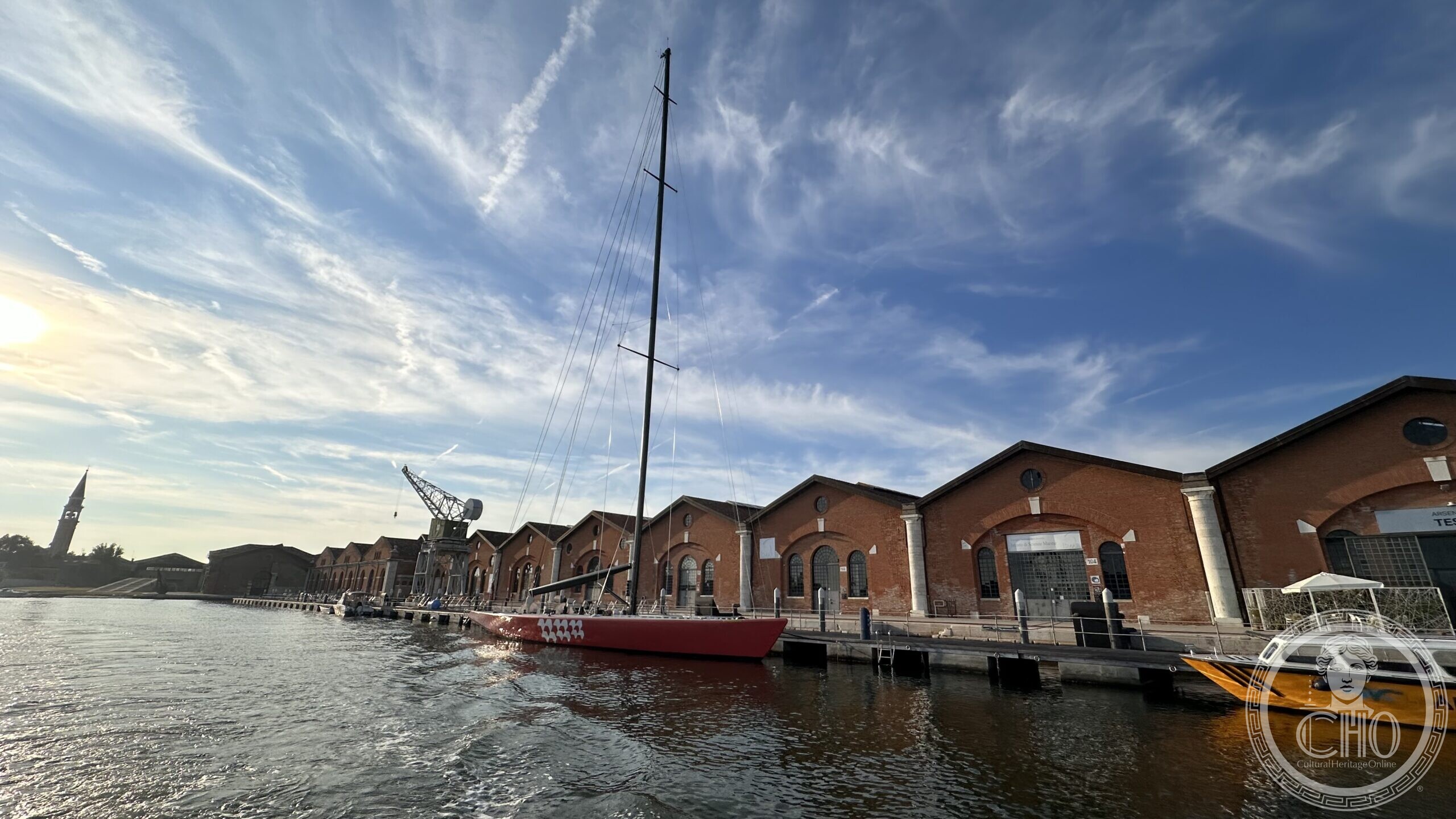 Wide view of the Il Moro di Venezia yacht moored at the Arsenale Nord, mast against a bright sky, with the brick sheds of the Novissima dock alongside