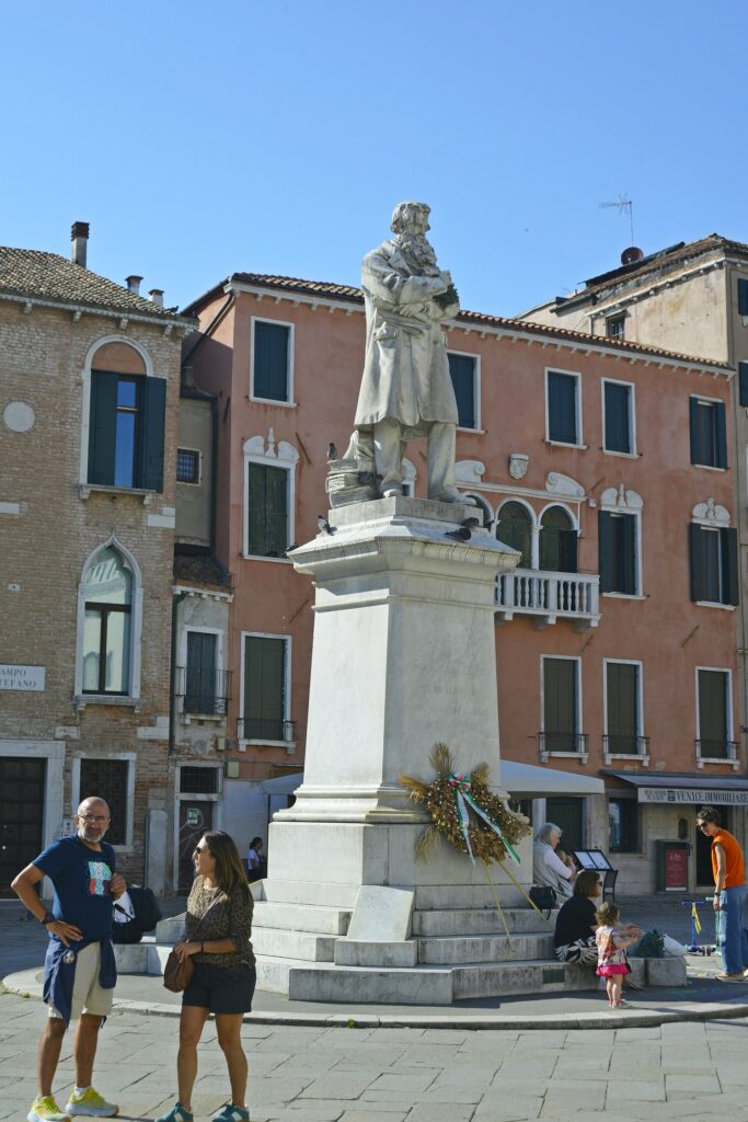A marble statue of a bearded man in a long coat on a high plinth, a stack of books showing under the coat